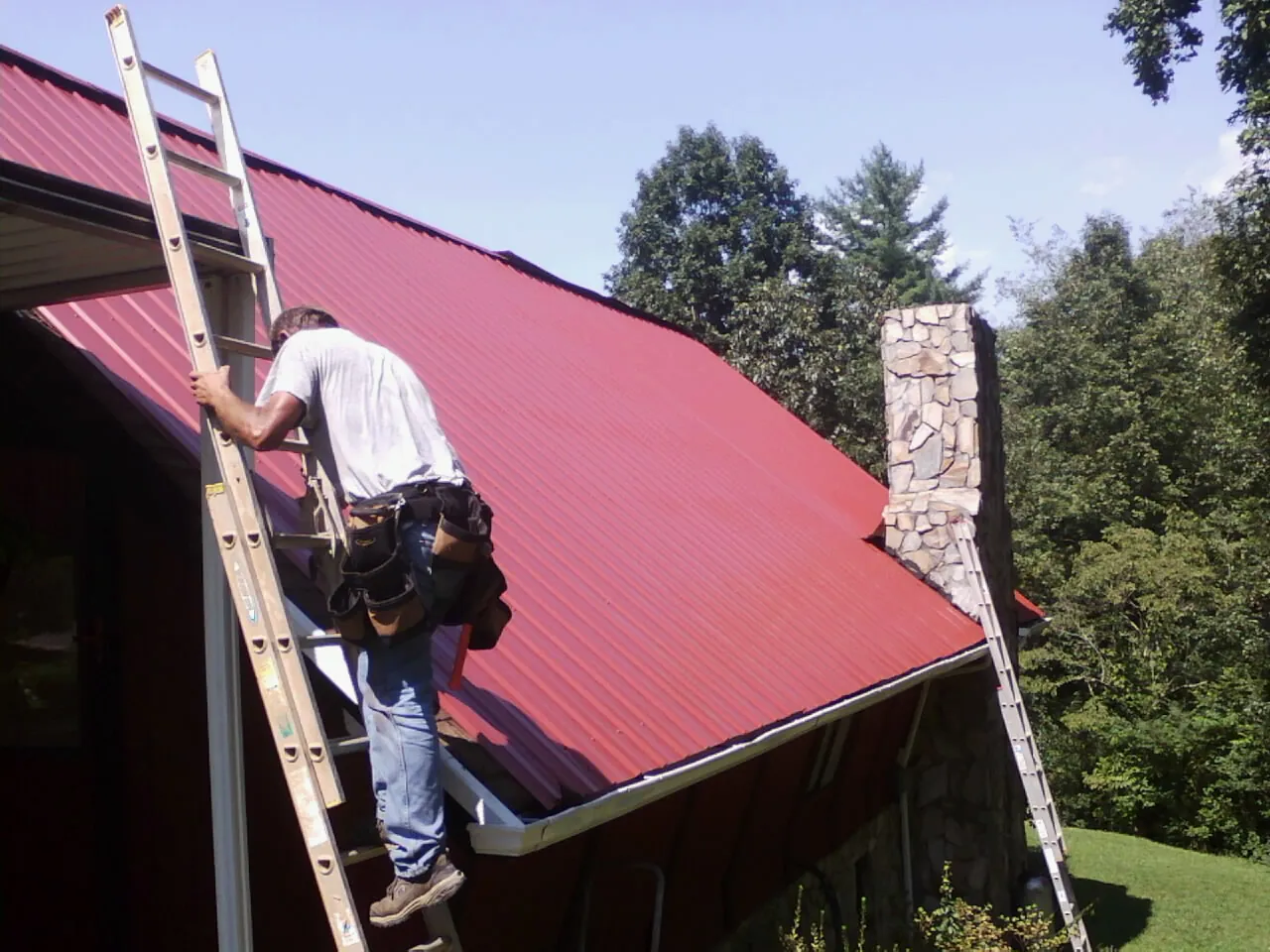 Roofer on a ladder beside a red metal roof and stone chimney — Soffit & Fascia Repair by Roofing Brothers in Bonne Terre